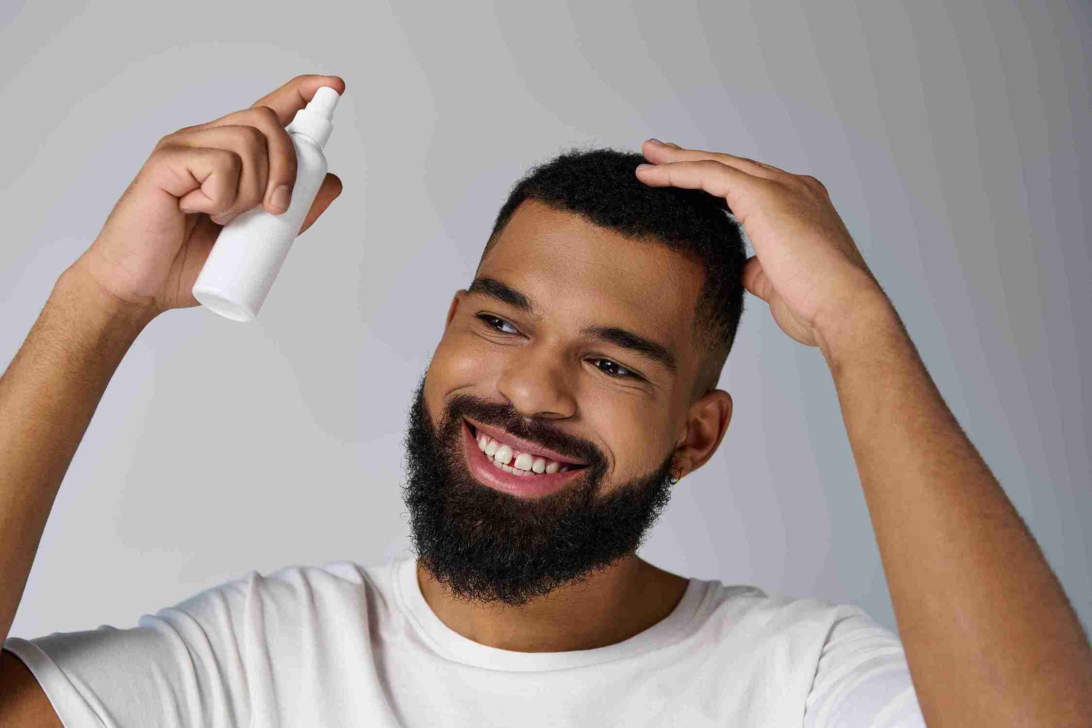 Lächelnder Mann mit Bart trägt weißes T-Shirt und sprüht Haarwuchsmittel auf seinen Kopf.